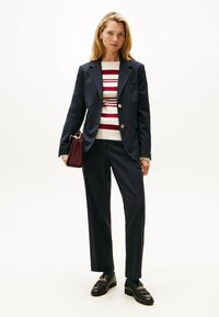 Blazer blu navale su misura con bottoni dorati, maglione rigato crema e rosso, pantaloni blu abbinati, mocassini neri con dettagli in catena, borsa bordeaux.