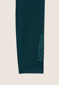 Leggings aderenti di un verde petrolio scuro realizzati in tessuto elasticizzato. Presentano il logo "FREDDY" in una tonalità più chiara lungo il lato. Cuciture piatte.