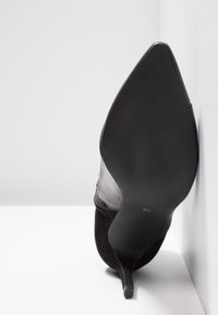 Escarpin noir à bout pointu avec semelle texturée et talon aiguille fin, positionné droit sur un fond blanc.