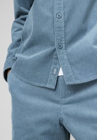 Chemise en velours côtelé bleu à rayures verticales, avec fermeture par boutons et une étiquette portant l'inscription "BZB". Associée à un pantalon en velours côtelé assorti.