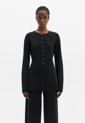 Femme portant un top noir ajusté à manches longues boutonné et un pantalon large assorti, debout devant un fond blanc uni.