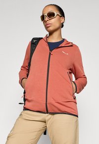 Κόκκινο-πορτοκαλί φλιτζάνι με κουκούλα, μαύρο φερμουάρ και λογότυπο. Συνδυάζεται με μπεζ παντελόνι, με υφασμάτινη επιφάνεια και τσέπες.