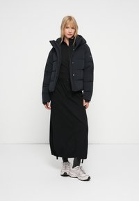 Kvinna klädd i svart puffjacka, svart lång kjol med cargofickor, grå strumpor och ljusgrå sneakers, stående mot en neutral bakgrund.
