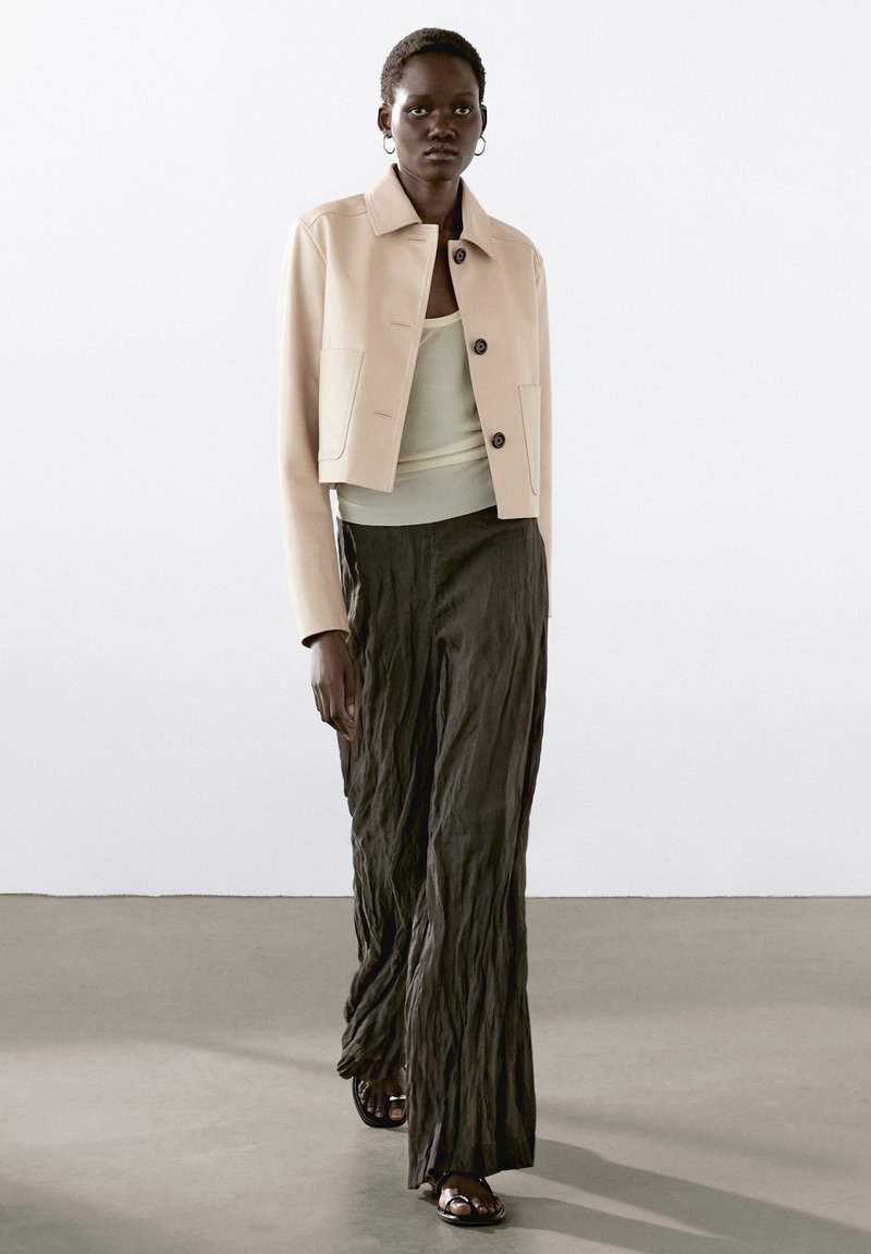 Beige cropped jakke med knapper foran, parret med en lys grøn tanktop og strukturerede mørke bukser. Modellen bærer flade sandaler.