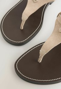 Paire de sandales en cuir marron avec des brides en daim beige en forme de lanière et une couture blanche autour des bords de la semelle, posées sur une surface claire.