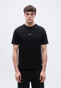 Νεαρός άνδρας με σγουρά μαλλιά  φοράει εφαρμοστή μαύρη μπλούζα με το κείμενο «Boss Club» στο κέντρο του στήθους, σε απλό λευκό φόντο.