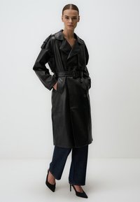 Cappotto trench in pelle nera con fronte doppiopetto, ampi revers, vita con cintura e due tasche laterali. Abbinato a pantaloni in denim scuro.