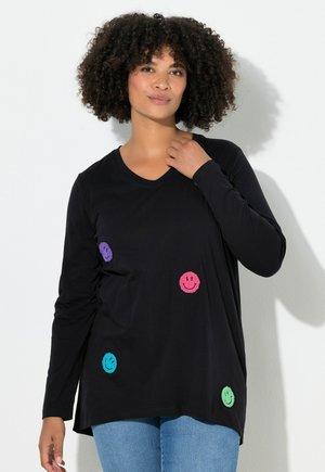 Nainen, jolla on kihara hiukset, pukeutunut mustaan pitkähihaiseen paitaan, jossa on värikkäitä brodeerattuja hymyileviä naamoja, sekä sinisiin farkkuihin, seisoo valkoista seinää vasten.