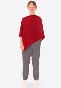 Poncho rosso lavorato a maglia con orlo asimmetrico, abbinato a pantaloni grigi a taglio dritto e sneakers bianche. Lo sfondo è bianco semplice.