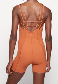 Femme portant une combinaison short sans manches de couleur rouille avec plusieurs fines bretelles et un petit logo Nike dans le haut du dos.