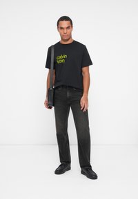 Vīrietis ģērbjas melnā Calvin Klein t-kreklā, tumšās džinsās, melnās kurpēs un nēsā melnu pleca somu, stāvot uz baltas fona.