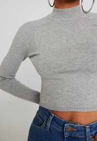 Suéter corto de punto acanalado gris con un cuello alto y mangas largas ajustadas, combinado con jeans de mezclilla azules.