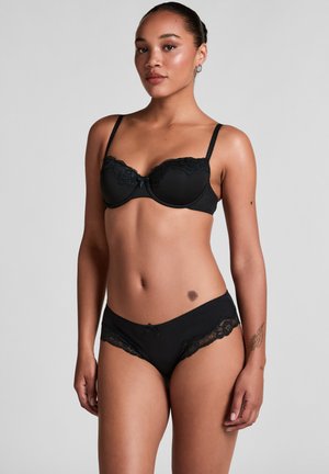 Vrouw staand, dragend een bijpassende zwarte kanten beha en slip, met zichtbare tatoeages op de linker onderarm en rechter heup.