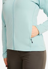 Person som bär en ljusblå zip-jacka med handen i den dragkedjeförsedda fickan och mörkbruna byxor med en dragkedjeförsedd sidficka.