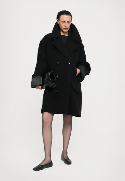 Black double-breasted coat with a wide collar, oversized sleeves, and a soft texture. Model holds a black clutch; paired with black flats and sheer tights.