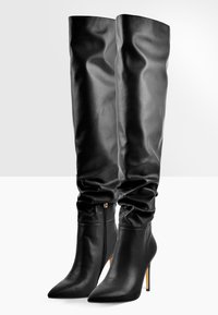 Bottes en cuir noires montant jusqu'aux genoux, à bouts pointus, tiges froissées, fermetures éclair sur les côtés et talons aiguilles sur fond blanc.