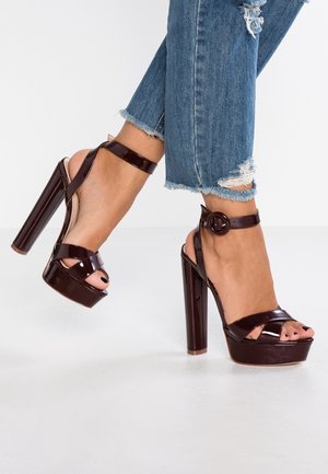 Sandały na wysokim platformowym obcasie w ciemnym bordowym lakierowanym skórze. Charakteryzują się krzyżującym się wzorem, paskiem wokół kostki i ozdobnym okrągłym klamrą.