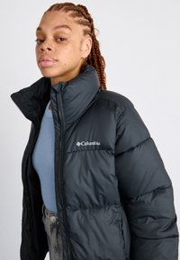 Giacca a vento nera con colletto alto, dotata di chiusura con zip e logo Columbia sul lato sinistro del petto. Indossata sopra una maglietta blu.