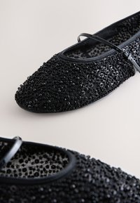 Ballerines Mary Jane noires avec des embellissements en perles et une fine bride sur le cou-de-pied, sur un fond clair et neutre.