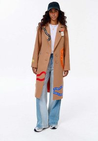 Žena v béžovém kabátu s barevnými nášivkami "NEZASTAVUJ", světle modrých širokých džínách, bílých teniskách, bílém tričku a černé baseballové kšiltovce.