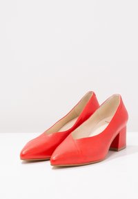 Ein Paar rote Leder-Pumps für Damen mit niedrigem Absatz und spitzen Zehen, beigem Innenfutter und Blockabsätzen auf einer weißen Oberfläche.