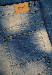 Blue faded denim jeans with back pocket and brown leather brand label reading "Reell est. 1997" stitched on waistband.