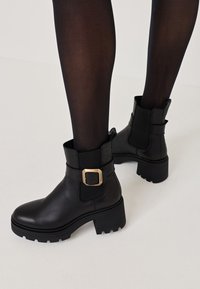 Bottes noires en cuir à la cheville avec un talon épais, des panneaux élastiques sur les côtés et un détail en boucle dorée sur la sangle. Semelle en caoutchouc texturée.