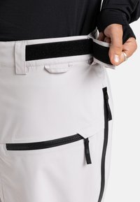Lichtgrijze broek heeft een klittenbandsluiting in de taille, zwarte ritszakken en een strakke, gladde textuur. Het ontwerp omvat minimale stiksels.