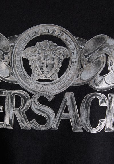 Tissu noir avec une inscription audacieuse en lettres argentées épelant "VERSACE" et un emblème de Méduse entouré d'un design de chaîne décoratif.