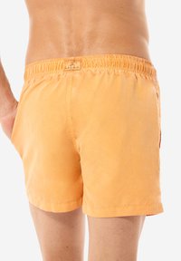 Costumi da bagno arancioni con vita elastica, texture del tessuto liscia e un'etichetta sul retro. Presentano un taglio classico con tasche laterali.