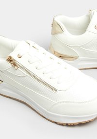Vita sneakers i texturerat läder, med dragkedjedetalj på sidan och metalliska accenter i guld. Har en gummisula och snörning framtill.