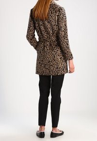 Abrigo largo de estampado de leopardo con textura suave, que presenta un corte entallado y mangas largas, combinado con pantalones ajustados negros y zapatos planos.