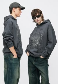 Sivé mikiny s kapucňou s grafickým dizajnom, nosené dvoma osobami. Mikiny majú voľný strih a rebrované manžety; kombinované s voľnými modro-zelenými džínsy.