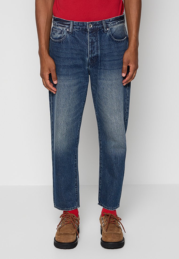 Osoba nosząca wyblakłe niebieskie dżinsy, brązowe buty na sznurówki i jasnoczerwone skarpetki, stojąca na tle jednolitego jasnego tła.