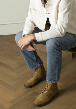 Leren veterschoenen van suède in lichtbruin met een ronde neus en rubberen zool, gecombineerd met een blauwe spijkerbroek en een witte jas, zittend op een houten stoel.