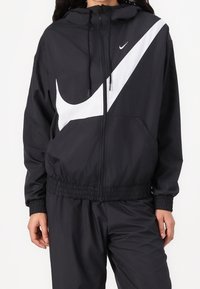 Svart vindjacka med en kontrasterande vit Nike-swoosh, elastiska muddar och midjeband, med blixtlåsförslutning framtill och huva.