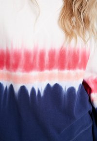 Chemise à manches longues tie-dye avec des bandes horizontales rouges, blanches et bleues portée par une personne aux cheveux blonds ondulés visibles à l'épaule.