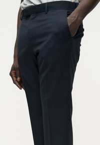 Homme portant un pantalon de costume bleu marine foncé, une main dans la poche, des passants de ceinture visibles, et une chemise gris clair rentrée.