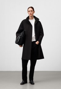 Cappotto oversize nero con colletto alto, camicia bianca sotto, pantaloni neri e scarpe nere. Borsa portata sotto il braccio, texture liscia.