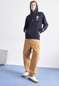 Hanorac bleu marin cu buzunar frontal și logo, asociat cu pantaloni cargo bej cu buzunare laterale și o croială confortabilă, completează ținuta cu teniși albi.