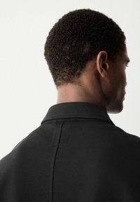 Chaqueta de color negro con un acabado texturizado, que presenta un diseño limpio y un corte en ángulo en los hombros. Enfoque en la vista posterior.