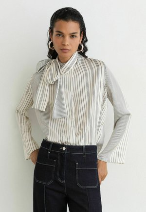 Kvinde iført en hvid bluse med sorte lodrette striber og en sløjfe i nakken, kombineret med mørke højtaljede jeans og store ringøreringe.