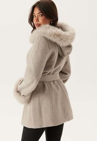Cappotto grigio chiaro con cappuccio e polsini in pelliccia sintetica, vita sottile e tessuto testurizzato. Presenta un design aderente e morbidi accenti di peluche.