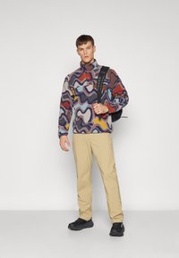 Fleece pullover dengan pola abstrak multicolor; celana beige, sepatu hitam; model memegang ransel hitam di satu bahu.
