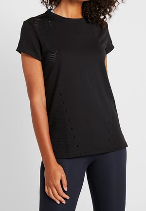 Femme portant un t-shirt de sport noir à manches courtes perforé et des leggings foncés, debout devant un fond clair uni.
