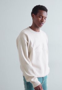 Un sweatshirt couleur crème avec une texture côtelée et un col rond, arborant un logo en relief subtil sur le devant. Associé à un jean clair.