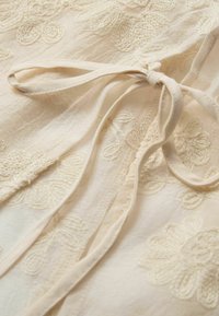 Tejido beige con bordados florales. Presenta un cierre de lazo y una superficie texturizada, que revela intrincados patrones de costura en toda su extensión.