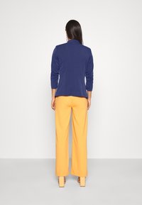 Veste bleu marine cintrée à manches longues associée à un pantalon large jaune vif. Textures lisses et arrière-plan neutre mettent en valeur la tenue.