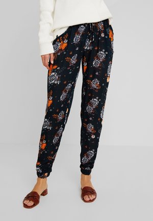Persona vistiendo pantalones jogger negros con patrones florales en naranja y blanco, combinados con tacones abiertos marrones y un suéter de punto blanco.