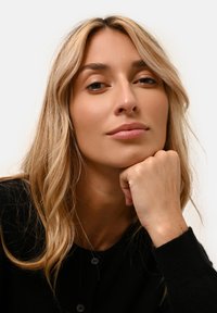 Blont hår med lösa vågor, neutraltonad hy, iförd en svart cardigan med knappar, vilar hakan på handen, framför en neutral bakgrund.